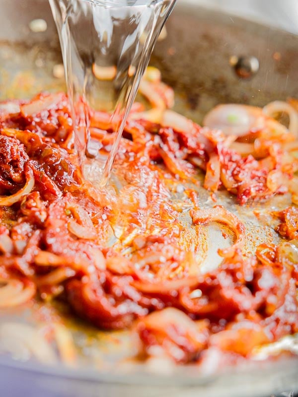 Close-up of a pan with tomato paste, sliced onions, and spices being mixed, while vodka is being poured in. The ingredients are sizzling, giving an impression of a cooking process.